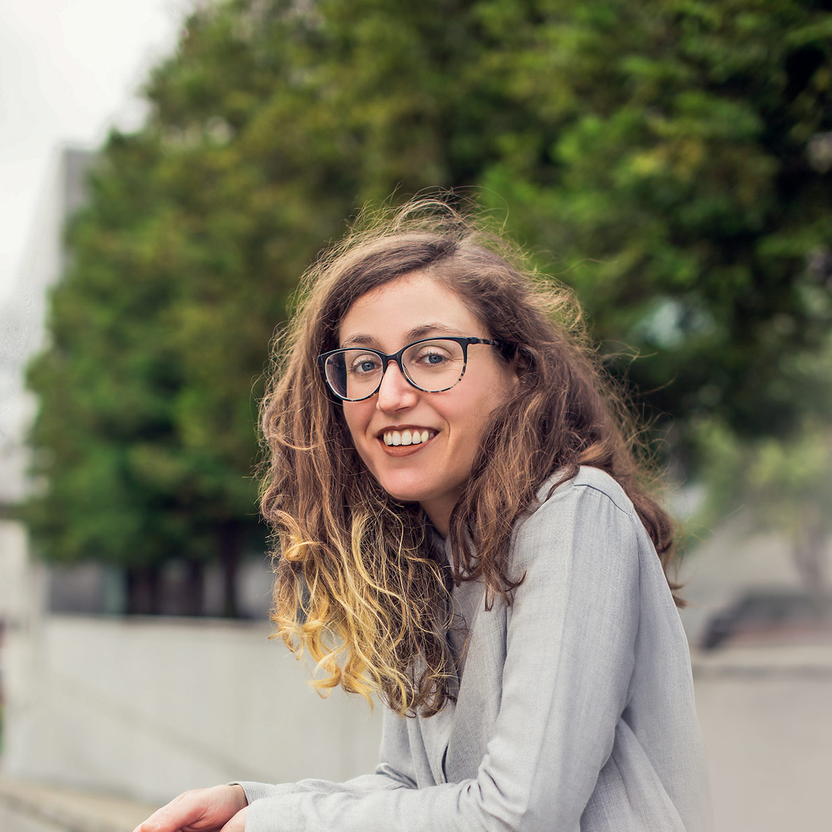 Howell stands leaning her elbows on a railing. She is smiling, wearing a gray button down shirt, with glasses.