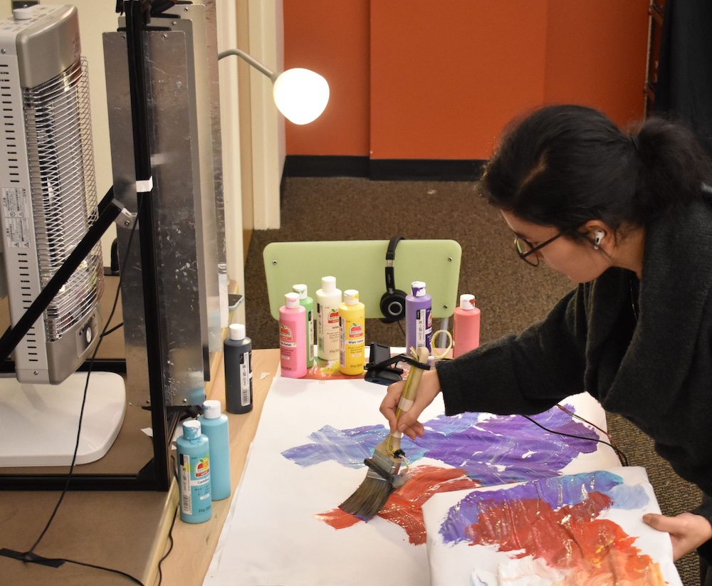 A person with glasses and dark hair leans over a flat canvas and paints a stroke of red. Across the table from them is a heater behind thermally insulating blinds.