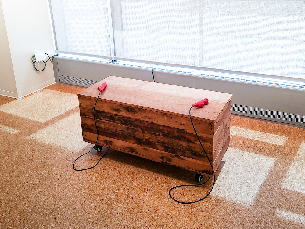 A wooden cedar chest has two red handles on top, with black cables coming out of the handles and curling around under the bench.
