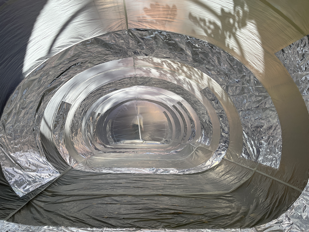 View from the inside of a large inflatable. Sheets of white and silver make up the side of a curvy tunnel. Light from the outside shines in.