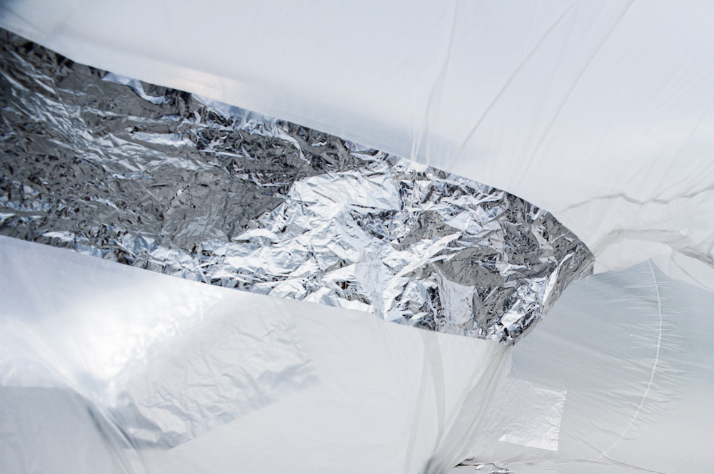 View from the inside of a large inflatable. Sheets of white and silver make up the side of a curvy tunnel. Light from the outside shines in.