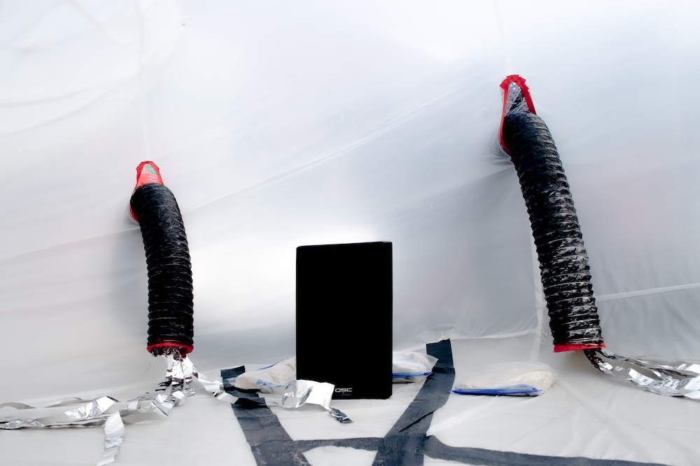 A black large speaker sits on the ground on the inside of the inflatable. On either side, black small air ducts enter the inflatable through holes cut in the side of the inflatable. The ducts reach down to the floor. Out of ducts come silver streamers. There is some black tape on the floor.