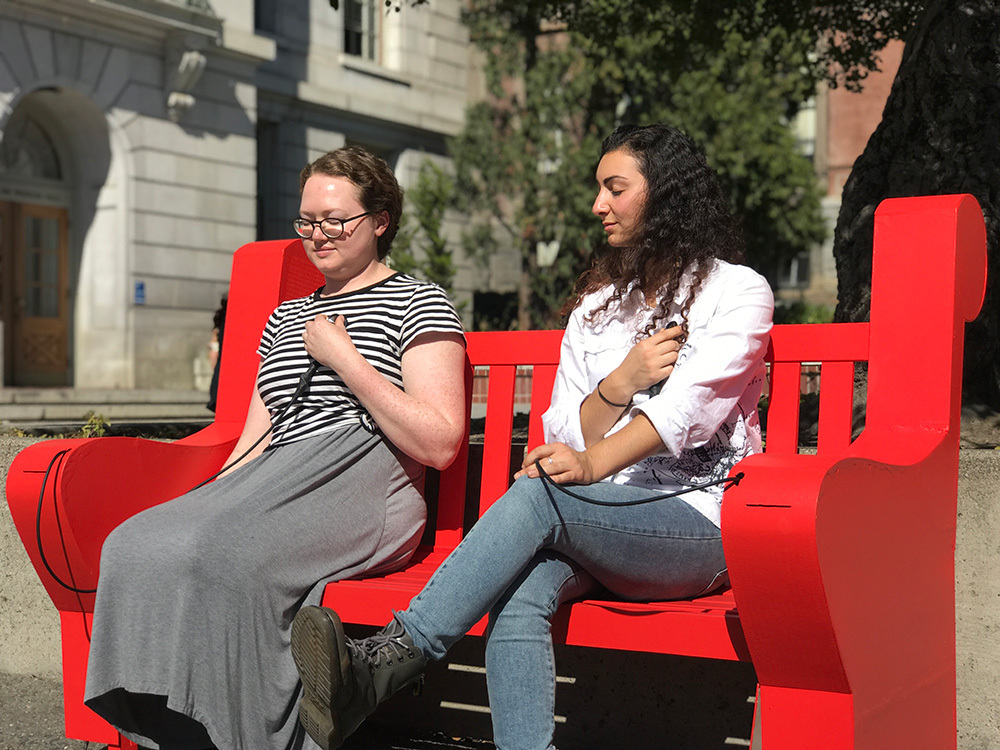 Two women sit on a red bench while holding stethoscopes to their chests. The stethoscopes end in long cables going into the bench. The women's eyes are closed and they look calm. The bench is outdoors. In the background is some foliage and a gray building.