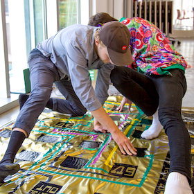 Two students demonstrate their Twister-like interface, stretching out and balancing on their limbs and hands on a mat on the floor. The floor map is gold with purple and green traces representing a circuit.