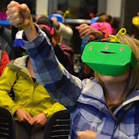 A crowd of people, whose faces are covered in bright paper masks that playfully look like VR headsets, are reaching their arms upward at slightly different angles.