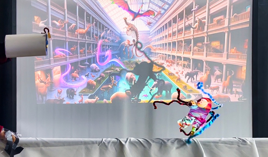 a puppet of paper and pipe cleaners is a unique weird playful creature performing in front of an AI-generated image projected on the back wall